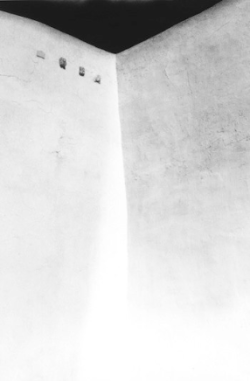 A high-contrast black and white photo of a bare corner wall with four small square holes near the top left.