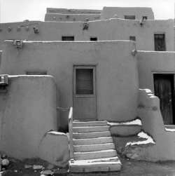 Adobe building with flat roofs, wooden doors, and a set of stairs leading to the entrance, partially covered in snow.