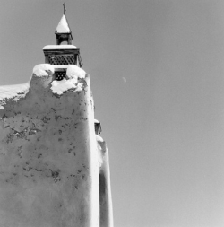 Snow covers the curved adobe wall and bell tower of a church; a faint moon is visible in the clear sky.