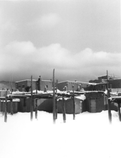 Adobe buildings with snow-covered roofs and wooden poles under a cloudy sky.