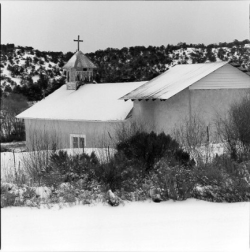 A snow-covered church with a cross on its steeple stands near bushes and trees in a winter landscape.