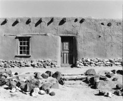 Adobe building with a single wooden door, one window, stone foundation, and rocks arranged in the sandy foreground.