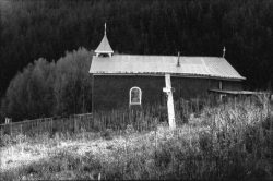 A small church with a metal roof stands on a grassy hill, with a forested area in the background.
