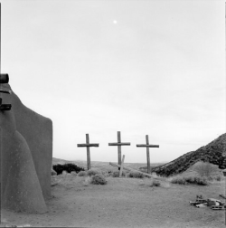 Three wooden crosses stand on a hill near an adobe building, with mountains and desert landscape in the background.