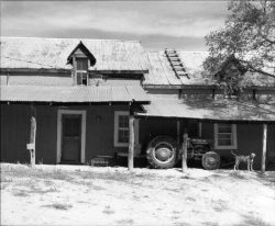 Black-and-white photo of an old farmhouse with a tractor, a wooden porch, and a dog standing near the front.