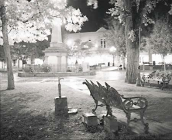 Black-and-white photo of empty benches near a monument in a well-lit park at night.