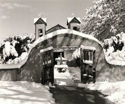 Snow-covered adobe church entrance with open wooden gates, revealing a courtyard and a cross in the background.