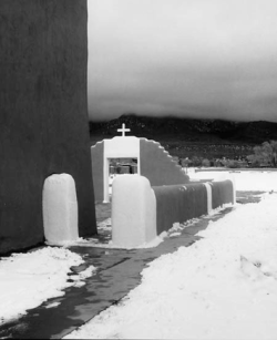 Snow surrounds an adobe church with a cross above its arched entrance, set against mountains and a cloudy sky.