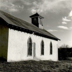 Small white church with arched windows and a steeple, photographed under a cloudy sky.