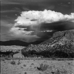 A small rural church and cemetery sit at the base of rocky hills under a dramatic, cloud-filled sky.