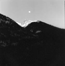The moon is visible above a snowy mountain peak, with dark forested hills in the foreground.