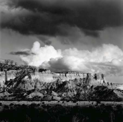 Black-and-white photo of dramatic clouds over a rocky mesa with flat-topped cliffs and sparse vegetation below.