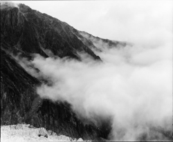 Black and white photo of mountains with low clouds or fog partially covering the rocky slopes.