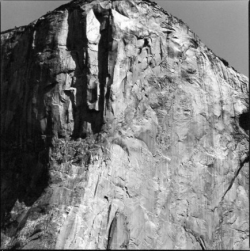 Black-and-white photo of a sheer, sunlit granite rock face with visible cracks and textured surface.