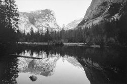 A calm lake reflects mountains, trees, and sky in a black-and-white landscape scene.