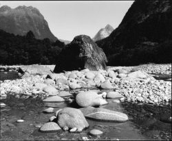 Large boulders and rocks line a shallow river with mountains and trees in the background under a clear sky.