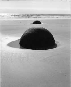 Two large, spherical boulders partially buried in wet sand on a beach near the shoreline under bright sunlight.