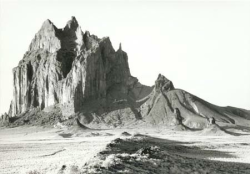 A large, jagged rock formation rises from a flat desert landscape under a clear sky.
