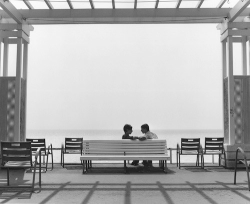 Two people sit and talk on a bench under a pergola, facing the ocean, with empty chairs around them.