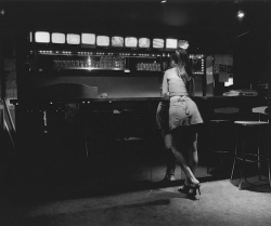A woman in a skirt and heels stands at a dimly lit bar counter with empty stools nearby.