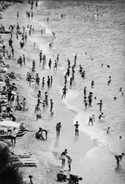Crowded beach scene with many people swimming, standing, and relaxing along the shoreline and in the water.