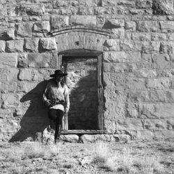 Person dressed in cowboy attire stands in front of a stone wall with a window opening, casting a shadow.