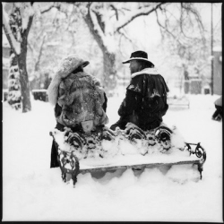 Two people in winter coats and hats sit on a snow-covered bench in a snowy park, facing each other.
