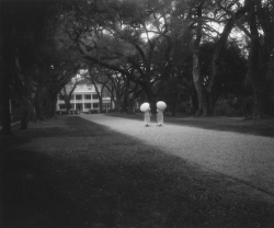 Two people holding parasols stand on a tree-lined path leading to a large house in the background.