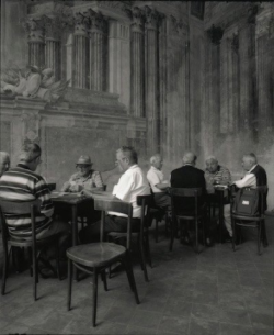Eight older men sit in two groups at tables, talking and playing cards in an ornate, historic-looking room.