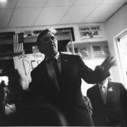 A man in a suit speaks to a group indoors, with posters and an American flag in the background.