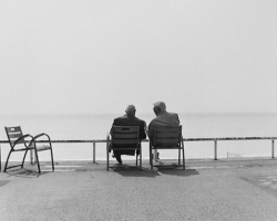 Two men sit on chairs facing the ocean, with empty chairs nearby and a clear sky above.