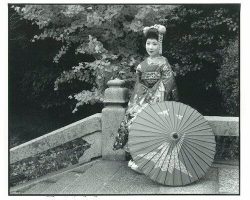 Woman in traditional Japanese attire stands on stone steps next to a paper parasol, with trees in the background.