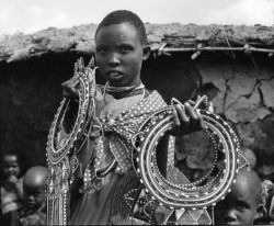 A person holds up two large, beaded necklaces while children stand nearby in front of a thatched-roof structure.