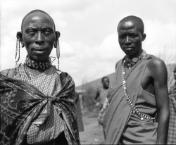 Two Maasai individuals in traditional attire and jewelry stand outdoors, with others visible in the blurred background.