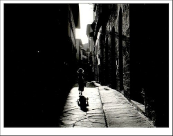 A silhouette of a child walking a dog down a narrow, sunlit alleyway with stone pavement.