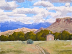 A small building stands in a grassy field with trees, mountains, and a cloudy sky in the background.