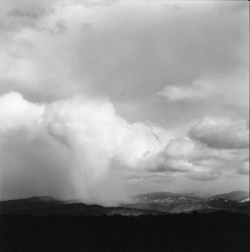 Black and white photo of clouds over distant mountains with rain falling from the sky above the landscape.