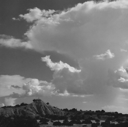 Black-and-white photo of a rocky hill with large, dramatic clouds in the sky above sparse vegetation.