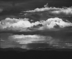 Dark clouds and streaks of rain hang over distant mountains in a dramatic, monochrome landscape scene.