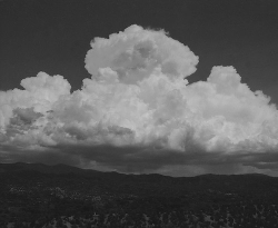Large, billowing clouds rise above a dark, hilly landscape under a mostly clear sky.