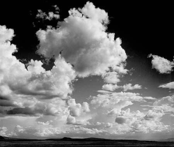 Black and white photo of large, dramatic clouds in a wide sky over a flat, open landscape.