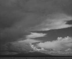 Black and white photo of dramatic clouds over a distant mountain range with dark water in the foreground.
