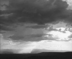 Black and white photo of sunbeams breaking through dark clouds over a distant mountainous landscape.