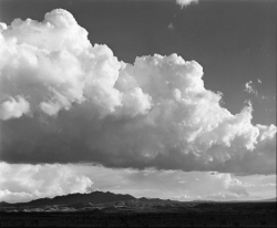 Large white clouds hover over distant mountains under a partly cloudy sky in a black and white landscape.