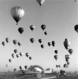 Black-and-white photo of multiple hot air balloons floating above buildings and a fenced area on a clear day.