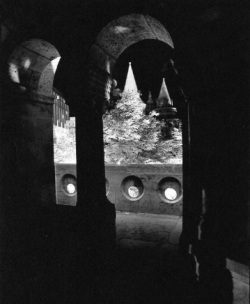 View through a dark arched stone passageway toward circular windows and pointed towers in the background.