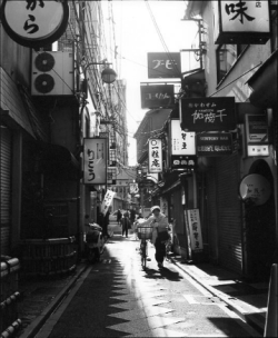 Narrow alleyway in a Japanese city with people walking and many signs in Japanese hanging above the street.