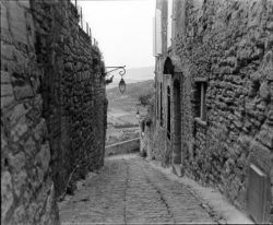 Narrow cobblestone alley lined with stone buildings, outdoor lamps, and a distant view of the countryside.