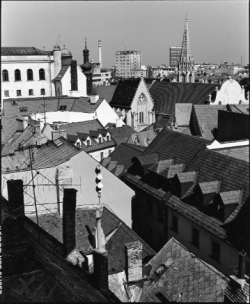 Black and white photo of a cityscape with tiled rooftops, church spires, and modern buildings in the background.