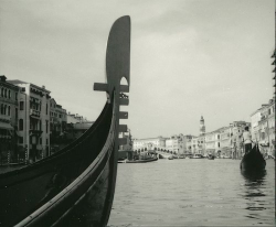 A gondola on a canal in Venice, with historic buildings and a distant bridge visible in the background.